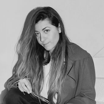 Black and white portrait of Federica Biasi in a dark coat, seated with a rectangular object, soft lighting, and a calm expression.