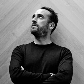 Man with beard and dark clothing stands against a diagonally paneled gray wall with wood grain