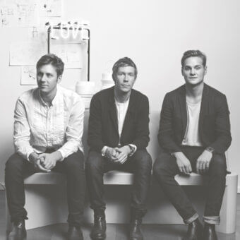 Black and white portrait of three men on a white bench, with handwritten wall art and a centered neon “LOVE” sign above.