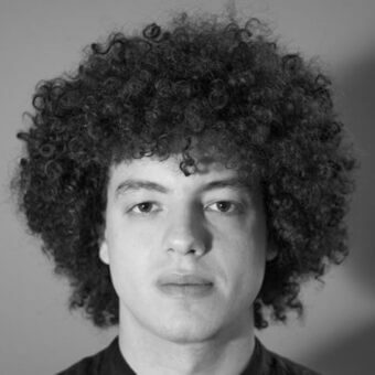 Black and white portrait of a man with a large afro hairstyle, dark eyes, and neutral expression against a gradient gray background.