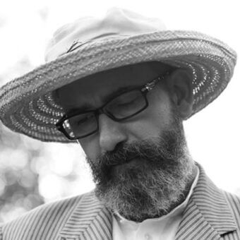 Man in straw hat and glasses, gray beard, striped blazer, contemplative pose, blurred foliage background, portrait by garattoni.