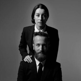 Two formally dressed people face forward, one with a hand on the other’s shoulder against a gray background