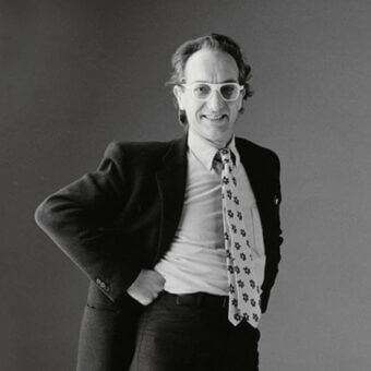Black and white portrait of Gaudenzi, a man with curly hair wearing a blazer, collared shirt, and floral patterned tie against a gray backdrop.