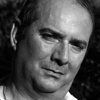 Monochrome portrait depicts a man facing slightly downward with dark hair, a beaded necklace, and shadowed facial features