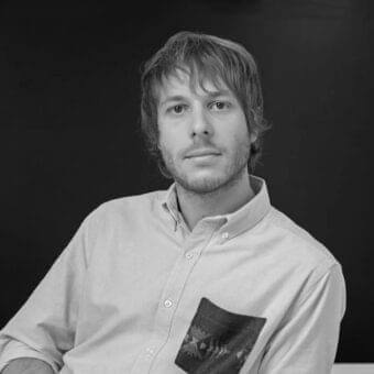Grayscale portrait of a man with layered hair and light facial hair wearing a partially unbuttoned button-down shirt against a dark gray background