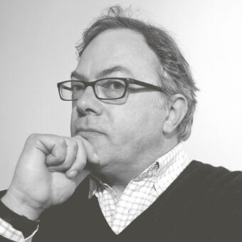 Gianni Rossetti portrait, gray hair, dark glasses, checkered shirt, dark sweater vest, contemplative pose, light gray background, diffused lighting.