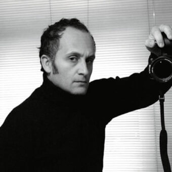 Black and white portrait of Giulio Mancini holding a digital camera with a black strap, against a backdrop of horizontal blinds.