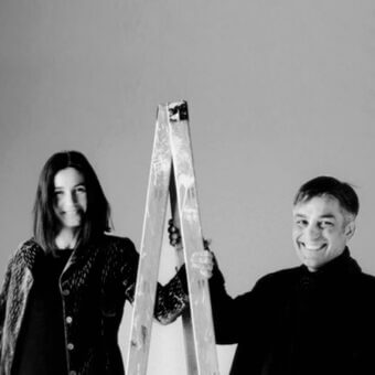 Black and white portrait of a couple with a wooden ladder; man holds ladder, woman gazes forward, plain gray background.