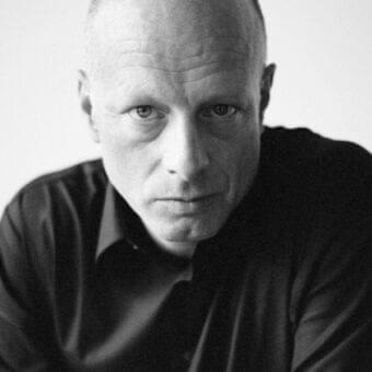 Light-skinned man with a hairless scalp and dark collared shirt looks directly at the camera with intense focus