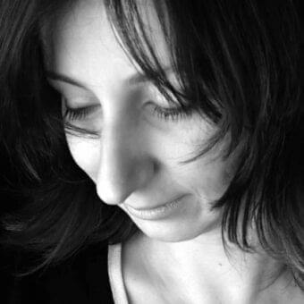Woman’s face in close-up with dark hair, downward gaze, and softly curved lips against a dark background
