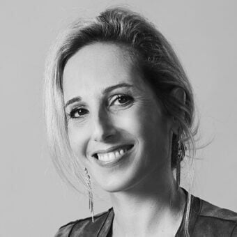 Black and white portrait of Irene Goldberg, fair skin, blonde updo, beaded earrings, warm smile, soft lighting, neutral gray background.