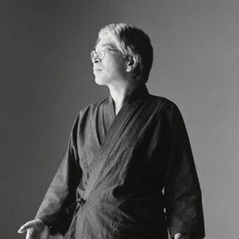 Black and white portrait of Isao Hosoe in dark kimono, wearing glasses, looking upwards with contemplative expression against gray background.