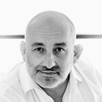 Black and white portrait of a man with shaved head, gray beard, wearing a light shirt, neutral expression, blurred background, soft lighting.