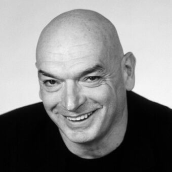 Black and white portrait of Jean Nouvel, a bald man in a black t-shirt, with a slight smile and tilted head against a white backdrop.