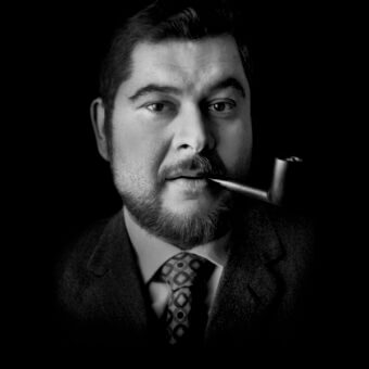 Black and white portrait of Joe Colombo in a tweed suit smoking a pipe, with mustache and beard, against a dark background.