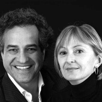 Two people face forward with gentle smiles, a man with curly hair and a woman with shoulder-length hair, against a dark backdrop