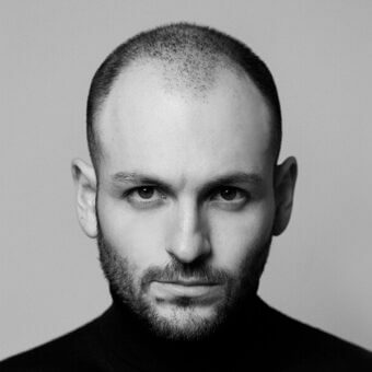 Black and white portrait of Leonardo Talarico, gray background, black turtleneck, close-up headshot, direct gaze, short beard, cropped hair.