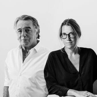 Two people, a man and a woman, face forward wearing eyeglasses and neutral-toned clothing against a pale backdrop