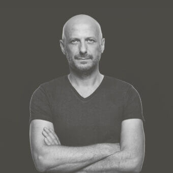 Black and white portrait of Luca Botto, shaved head, dark v-neck shirt, crossed arms, gray background, centered composition, formal presentation.