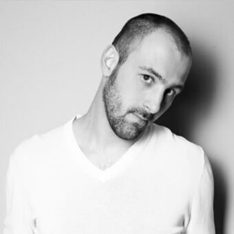 Black and white portrait of a man with short hair and beard wearing a white v-neck t-shirt, soft lighting, gray background.