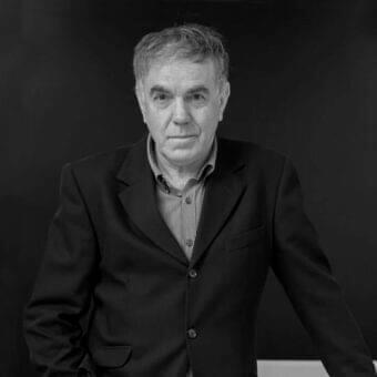 Gray scale portrait of a man with short hair wearing a dark jacket and striped shirt looking forward
