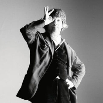 Black and white portrait of Luigi Serafini in velvet vest, collared shirt, and blazer, making a peace sign with raised hand.