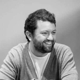 Grayscale portrait of a smiling man with curly hair and a beard wearing a V-neck and knitted cardigan