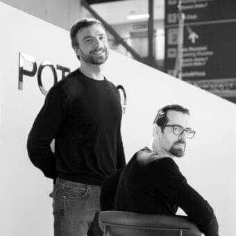 Two men stand and sit within a grayscale architectural space featuring angled walls with raised lettering and a dark staircase