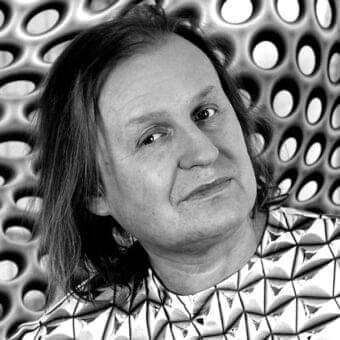 Person with layered hair wearing a patterned garment stands before a perforated wall in grayscale tones