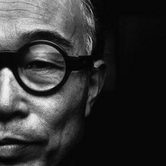 Grayscale close-up of a face with dark circular glasses, showing textured skin, wrinkles, and strong light creating shadows and contrast
