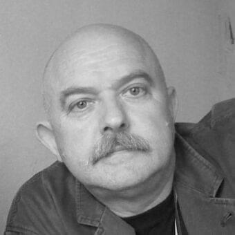 Bald man with a handlebar mustache looks forward wearing a dark jacket against a light wall