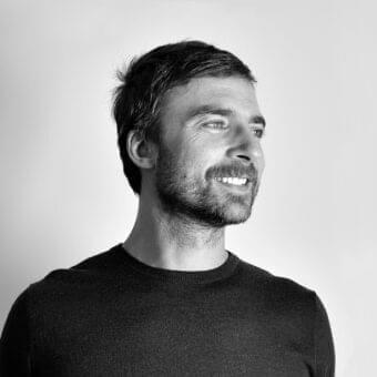 Grayscale portrait of a man with dark hair and beard wearing a dark round-neck garment against a light gray background