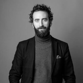 Man portrait, gray turtleneck and black blazer, curly dark hair, full beard, neutral expression, diffused lighting, gray background.