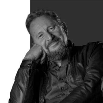 Grayscale portrait of a bearded man with hand raised, wearing a collared shirt and dark jacket against a divided background