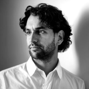 Black and white portrait of Mathias de Ferm, a man with curly hair and beard, wearing a white button-down shirt, gazing right.
