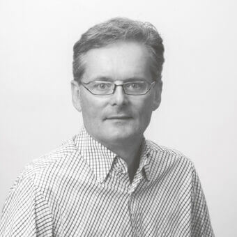 Portrait of Mauro Fadel, a man with short dark hair, wearing glasses and a light checkered long-sleeve shirt, against a gray backdrop.