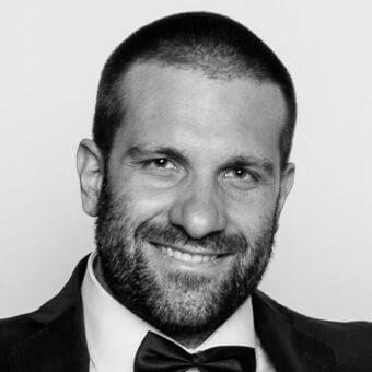 Black and white portrait of Michele Cazzaniga, a smiling man in a dark suit, white shirt, and black bow tie, with a graying beard.