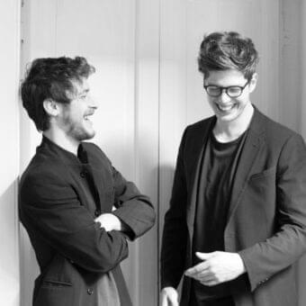 Two people in dark jackets stand close together before a light gray vertically paneled wall in grayscale