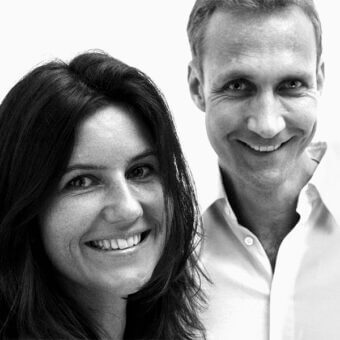 Black and white portrait of a woman and man smiling, both wearing collared shirts, against a light gray studio backdrop with “neuland industriedesign” text.