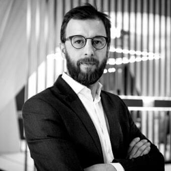 Grayscale portrait of a bearded man wearing glasses and a jacket with crossed arms looking forward against a blurred architectural background