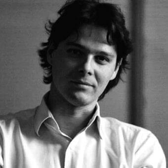 Young man with dark wavy hair looks toward camera wearing a light collared shirt against a blurred gradient background