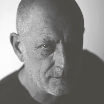 Bald man portrait, grayscale, dramatic lighting, shadowed eye, dark crew-neck shirt, textured skin, introspective expression, light gray background.