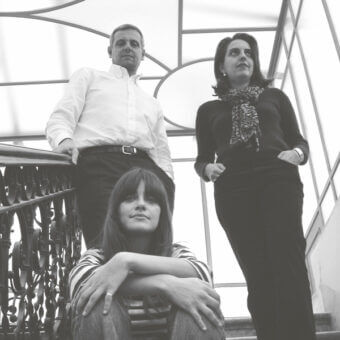 Black and white photograph of three people on a staircase in "pba studio," featuring a wrought-iron railing and geometric ceiling panels.