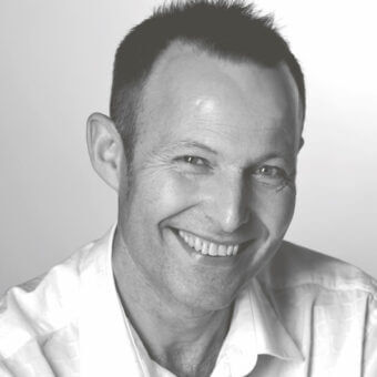 Black and white portrait of Peter Harvey, fair skin, short dark hair, light textured button-down shirt, soft gray gradient background.