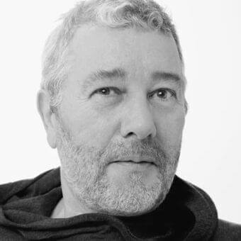 Grayscale portrait of a man with short gray hair and beard, wearing a dark hooded garment against a light neutral background