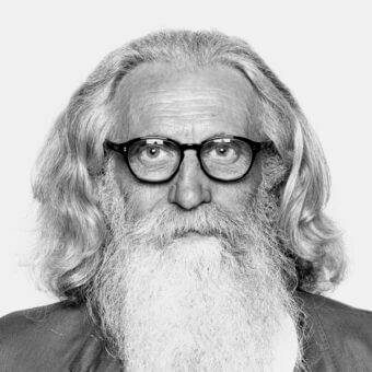 Black and white portrait of Piergiorgio Cazzaniga, a man with long silver-gray hair, beard, and round black eyeglasses, neutral expression.