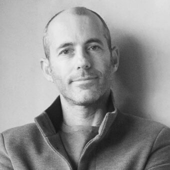 Black and white portrait of Pitsou Kedem, wearing a dark corduroy blazer with a stand-up collar, against a light gray wall.