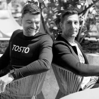 Two people seated in light chairs face away, wearing dark clothing and visible wrist accessories against a blurred outdoor backdrop
