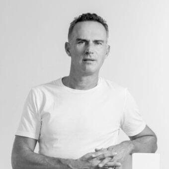 Fair-skinned man with graying hair looks forward wearing a white t-shirt with hands clasped against a light gray background