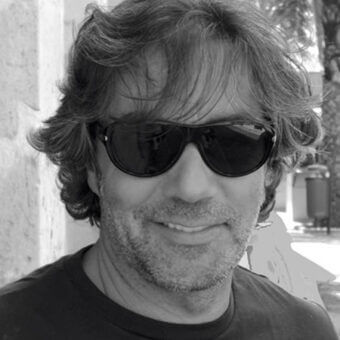Black and white portrait of Roberto Garbugli wearing dark rectangular sunglasses and a t-shirt, with wavy gray-streaked hair and a beard.